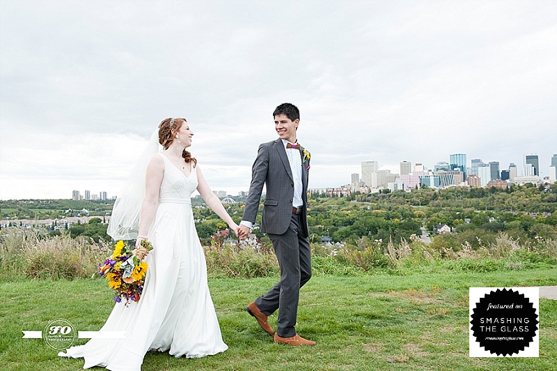 smiling married couple walking with Edmonton skyline in background, edmonton wedding photographers, bride holding bouquet, married couple holding hands and walking, 