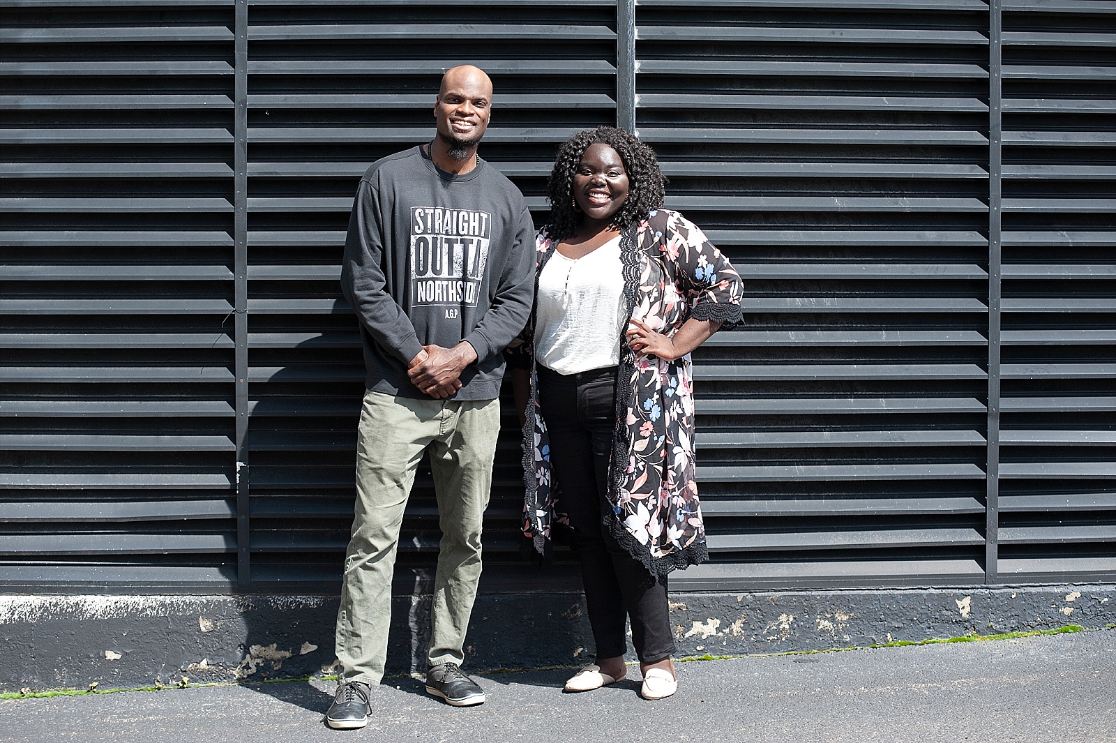 Smiling Black woman and Black man in front of black building vents.